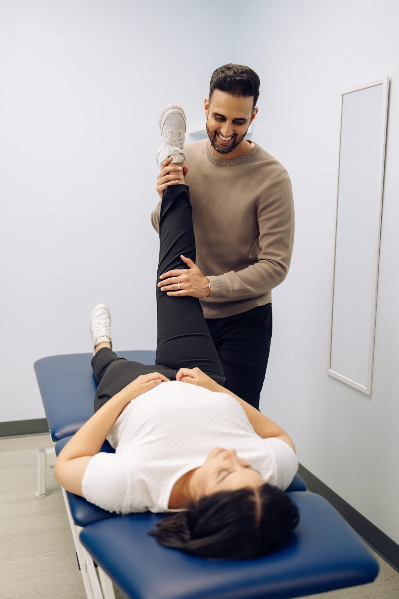 A man assists a woman with a leg stretch on a blue therapy table in a bright room.